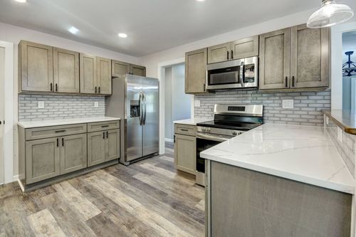 Modern kitchen with grey wooden cabinets, white marble countertops, a stainless steel refrigerator, and wood-look flooring.