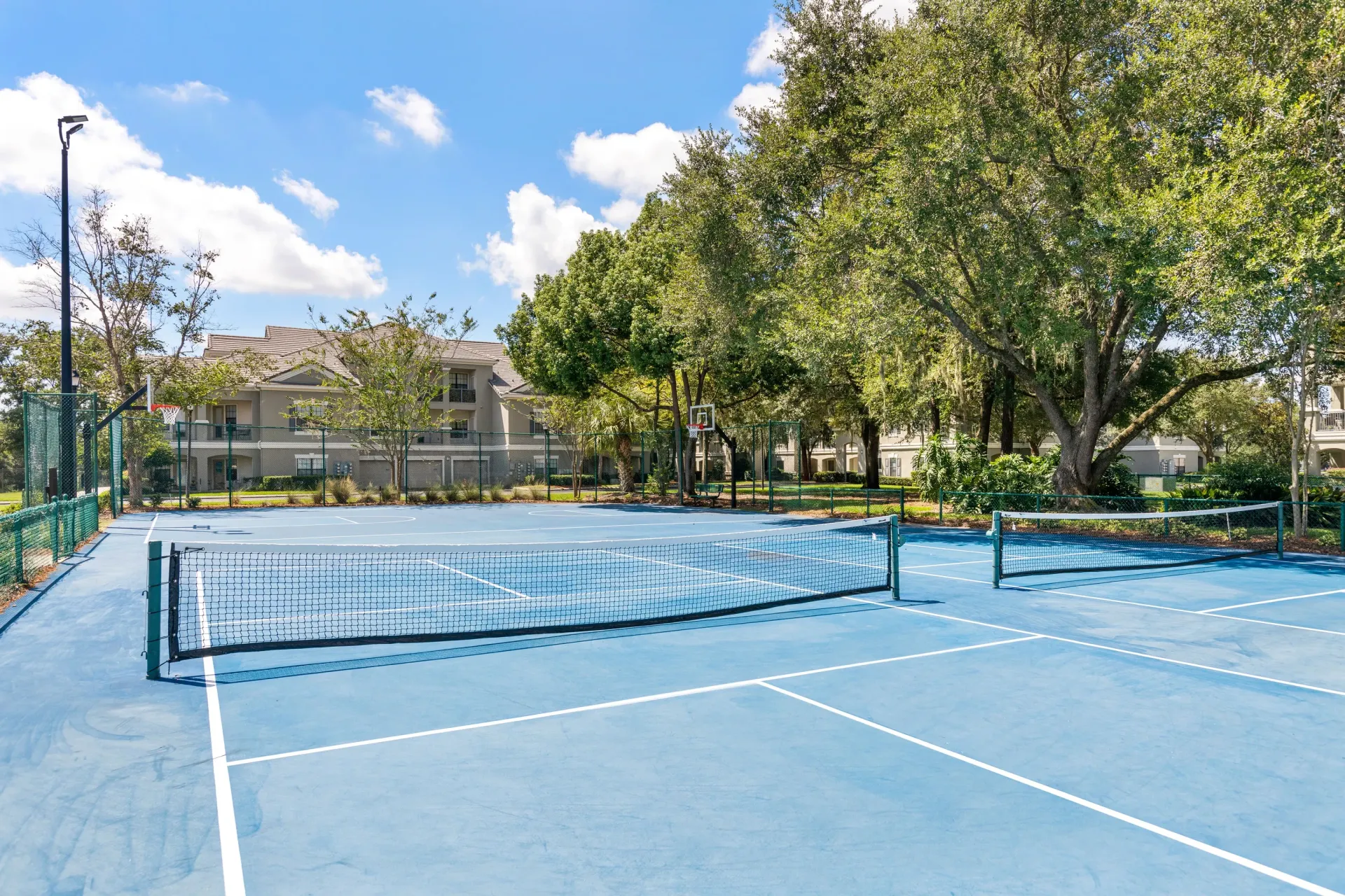 Empty outdoor tennis court with blue surface, net, trees, and a cloudy sky  at Levi at River City.