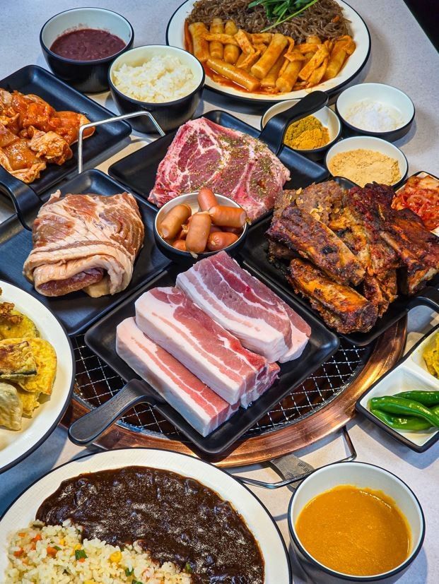 An abundant spread of BBQ dishes at Yukmijedang in Daegu, including raw meats ready to grill, rice, japchae, and various side dishes.