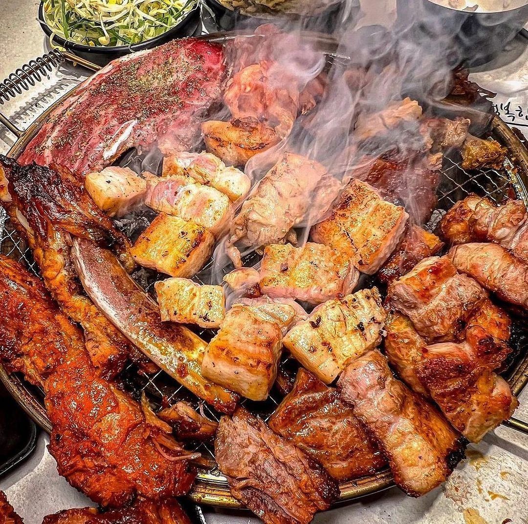 A mixed grill BBQ at Yukmijedang in Daegu, with a variety of meats including ribs, pork belly, and chicken cooking over an open flame.