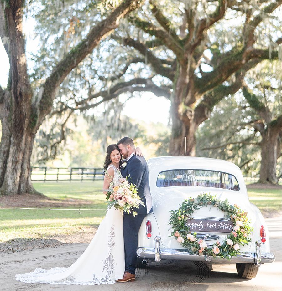 A bride and groom are standing next to a white car with a wreath on the back.