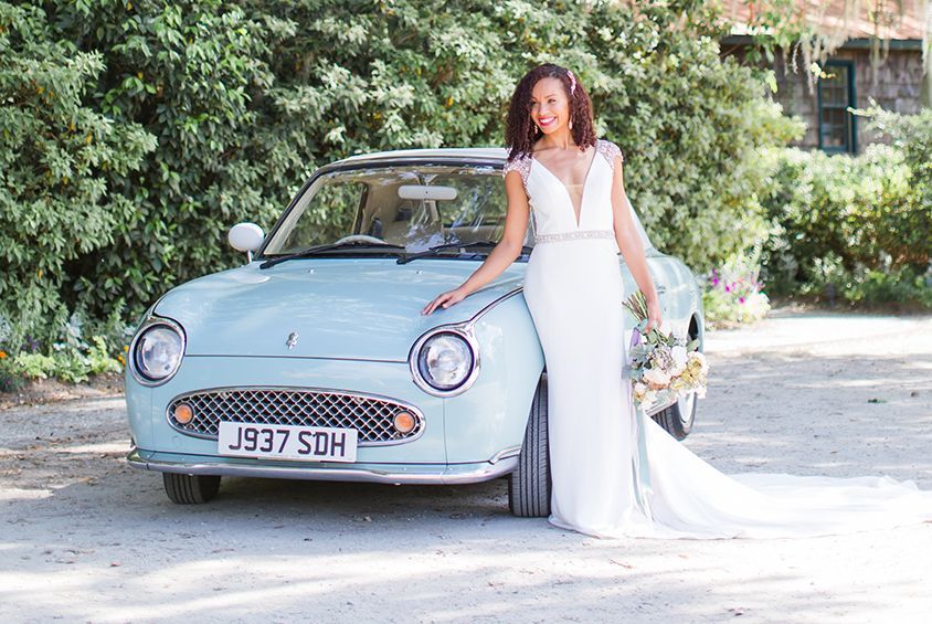 A bride in a white dress is standing next to a blue car.