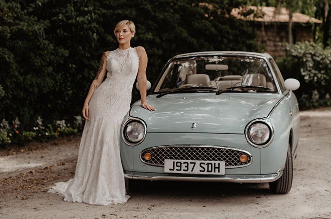 A woman in a wedding dress is standing next to a blue car.