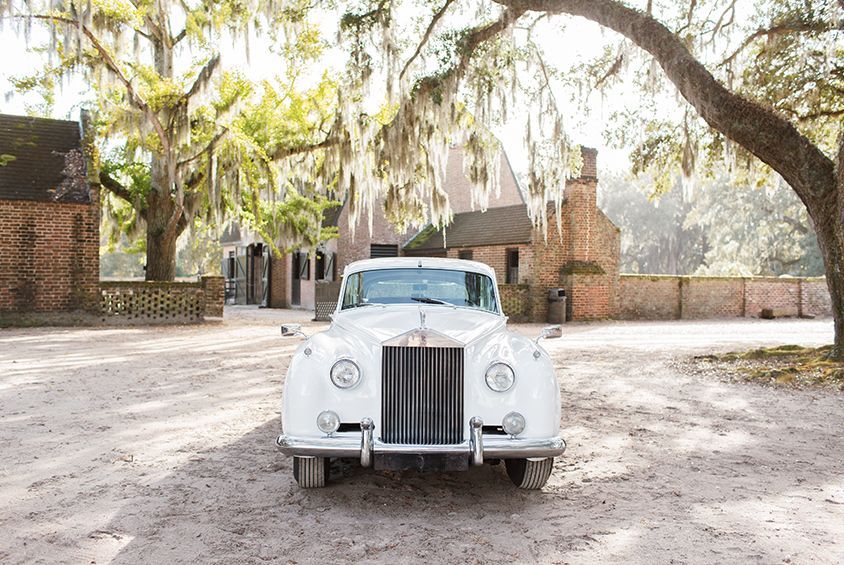 A white rolls royce is parked in a dirt lot in front of a building.