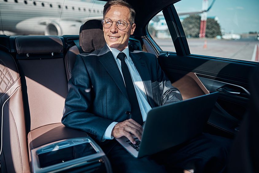 A man in a suit is sitting in the back seat of a car using a laptop computer.