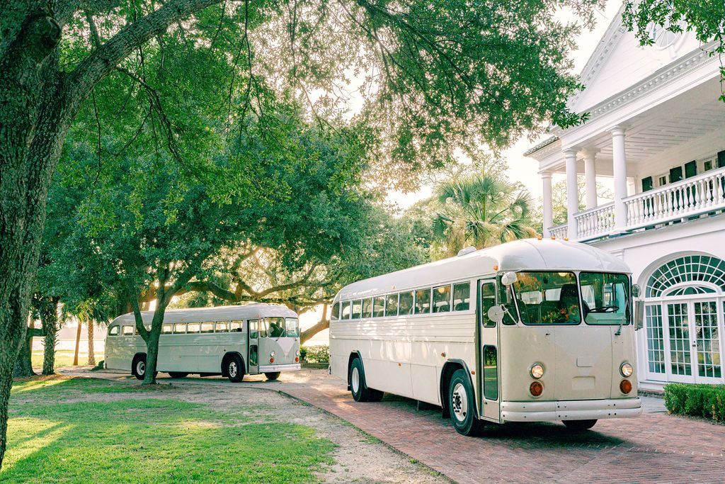 Two white buses are parked in front of a large white house.