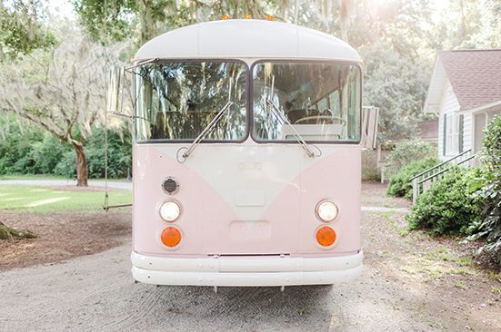 A pink and white bus is parked in a driveway in front of a house.