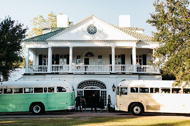 Two white buses are parked in front of a large white house.