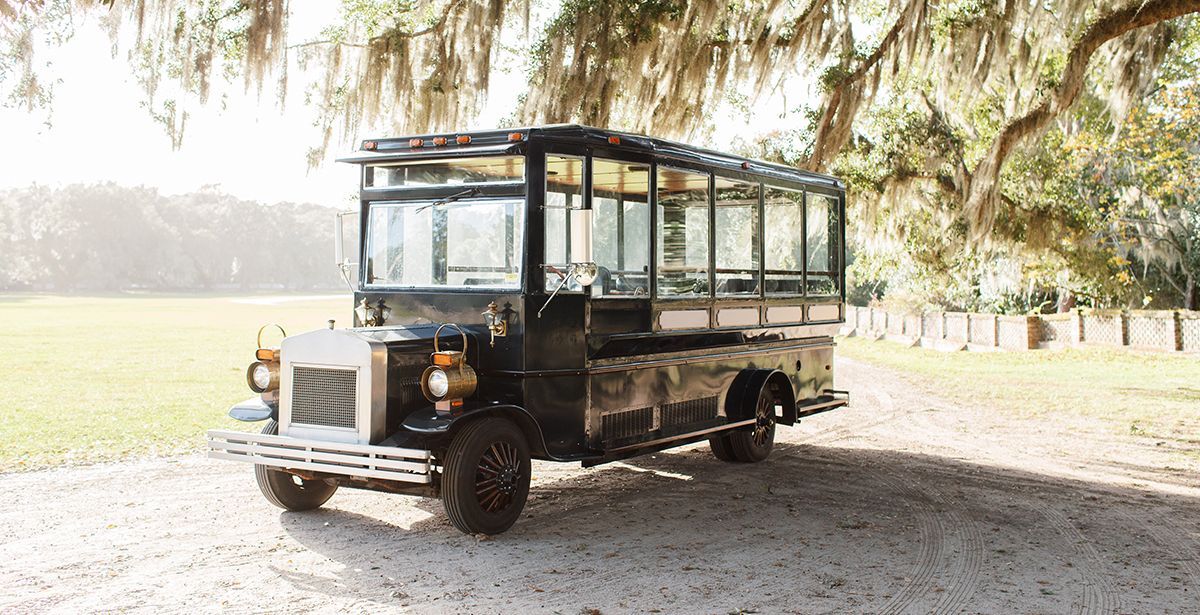 An old fashioned bus is parked in a gravel driveway next to a tree.