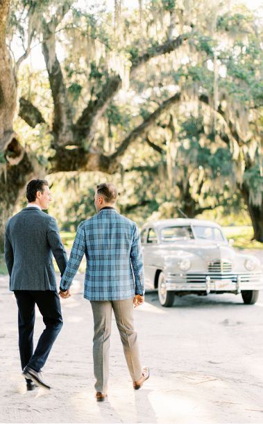 Two men are walking down a street holding hands in front of a white car.