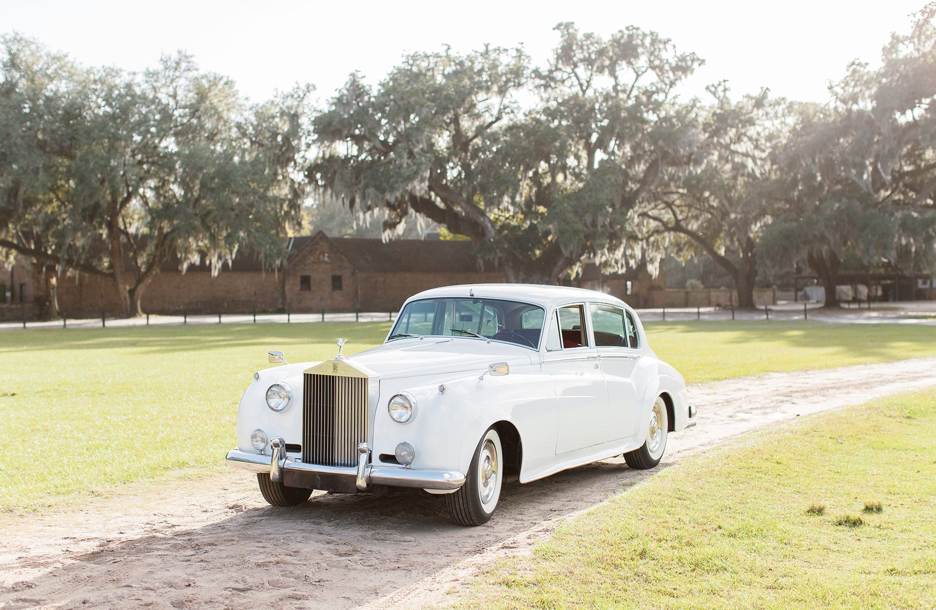 A white rolls royce is driving down a dirt road in a field.