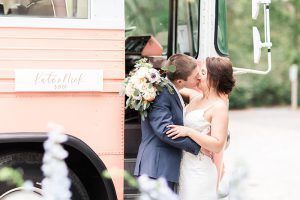 A bride and groom are kissing in front of a pink bus.