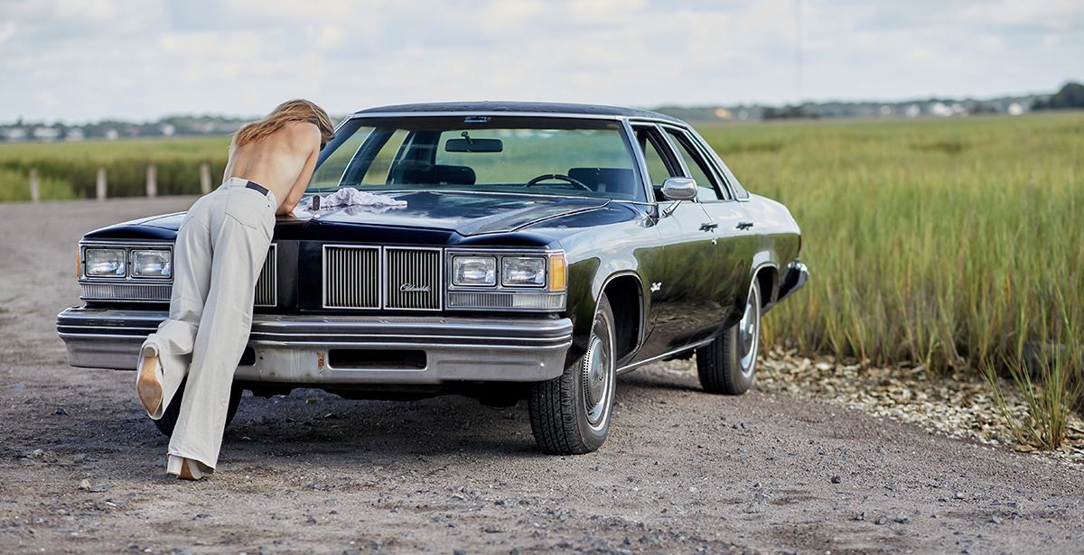 A woman is leaning against a car on a dirt road.