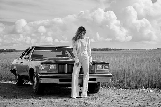 A woman in a white suit is standing next to a car in a field.