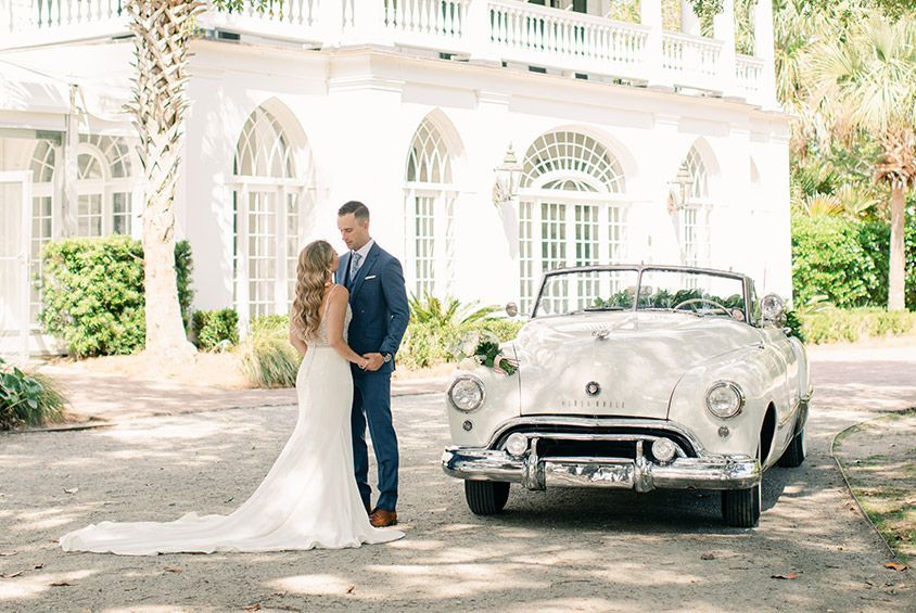 A bride and groom are kissing in front of a white convertible car.