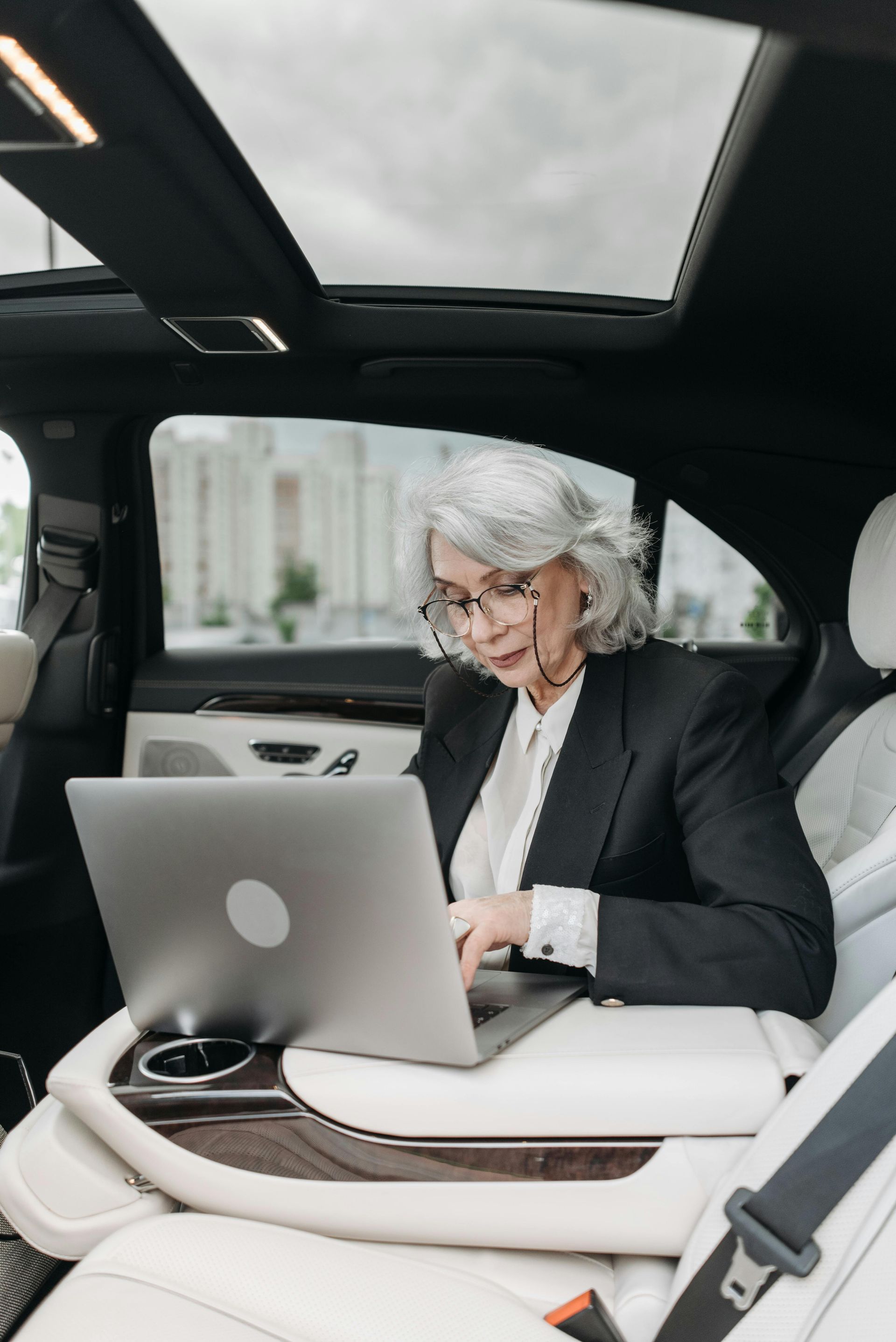 A woman is sitting in the back seat of a car using a laptop computer.