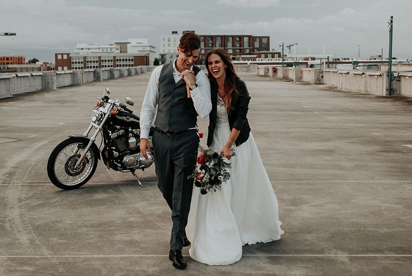 A bride and groom are walking in front of a motorcycle.