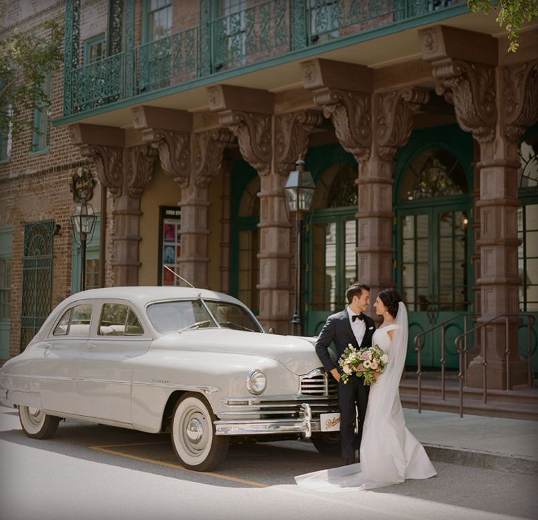A bride and groom are posing for a picture in front of a white car.