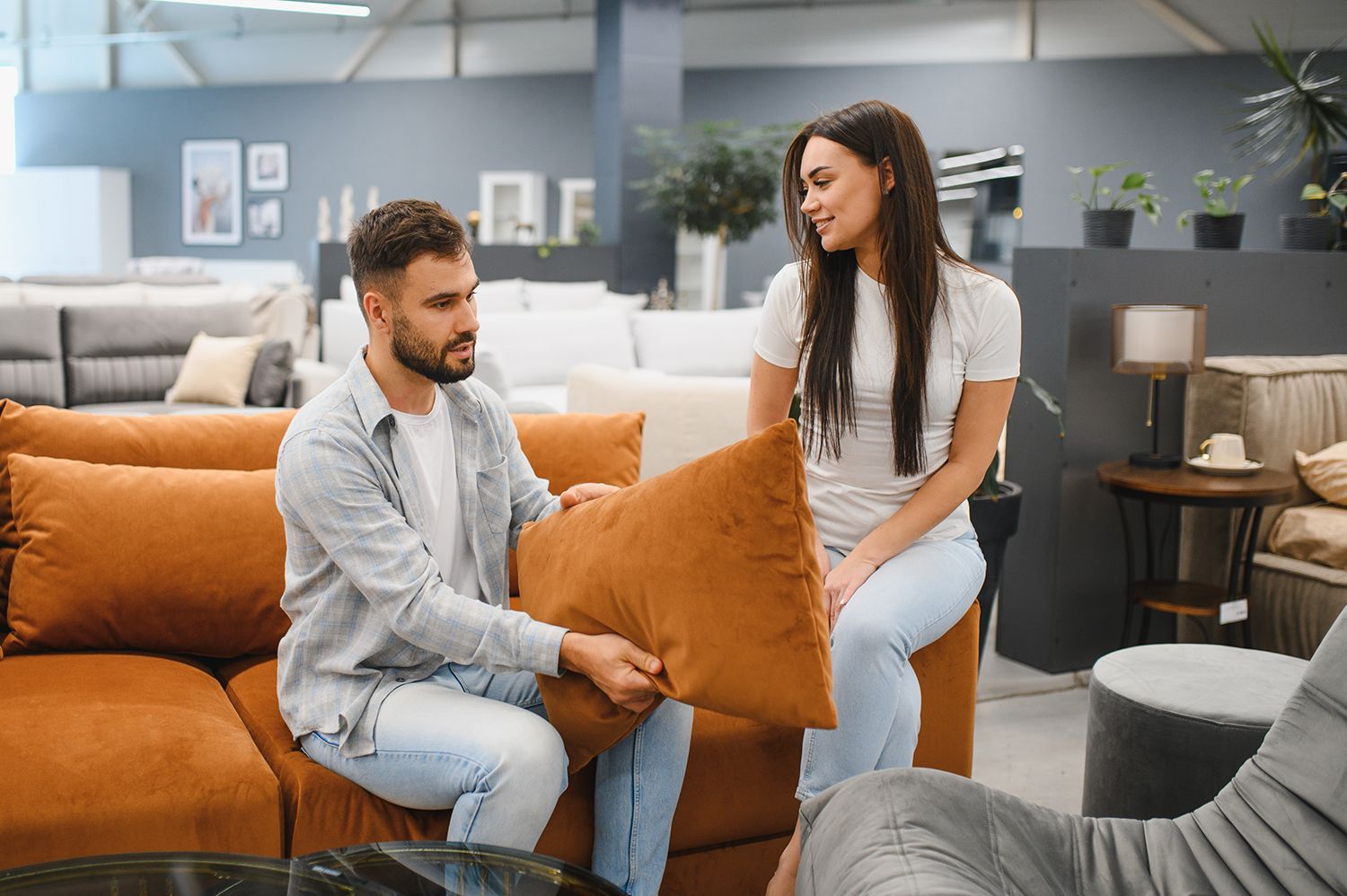 Two people sitting on a sofa in a furniture store, examining an orange throw pillow together.