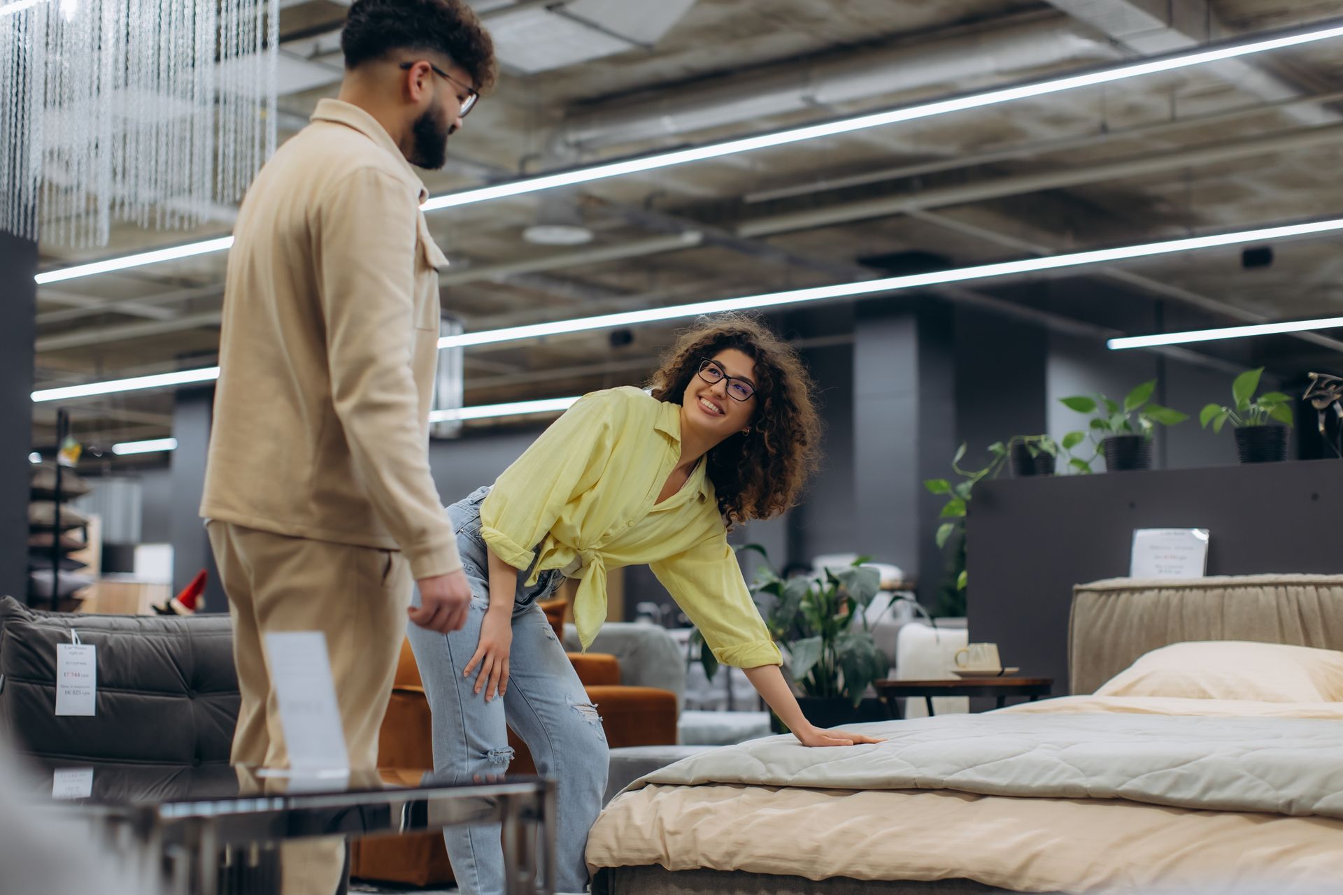 Two people testing a mattress in a furniture store showroom with beds and sofas.