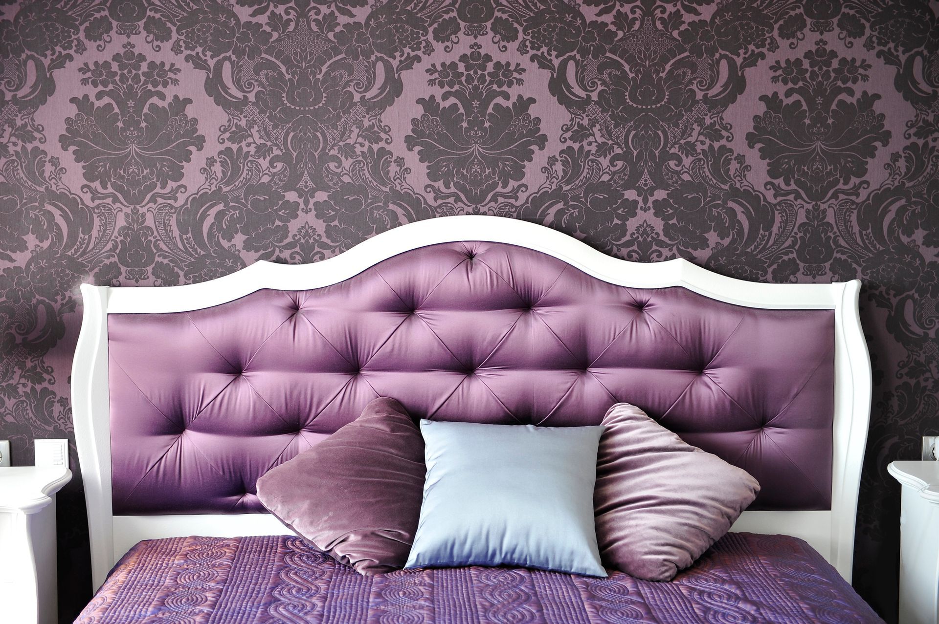 Luxurious purple bed with a white frame, with three multicolored small pillows on top.
