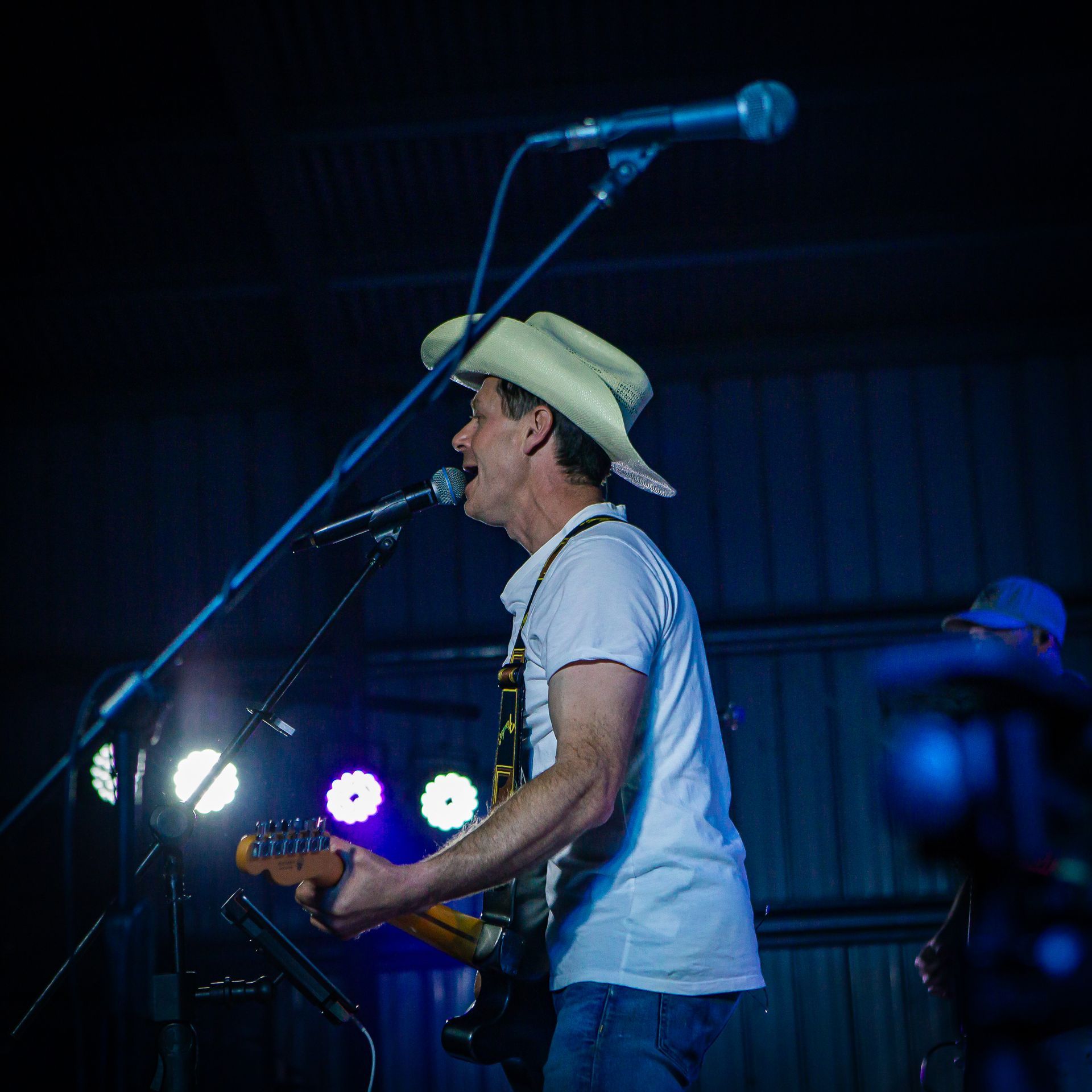 A man in a cowboy hat sings into a microphone while playing a guitar