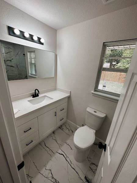 A bathroom with a toilet , sink , mirror and window.