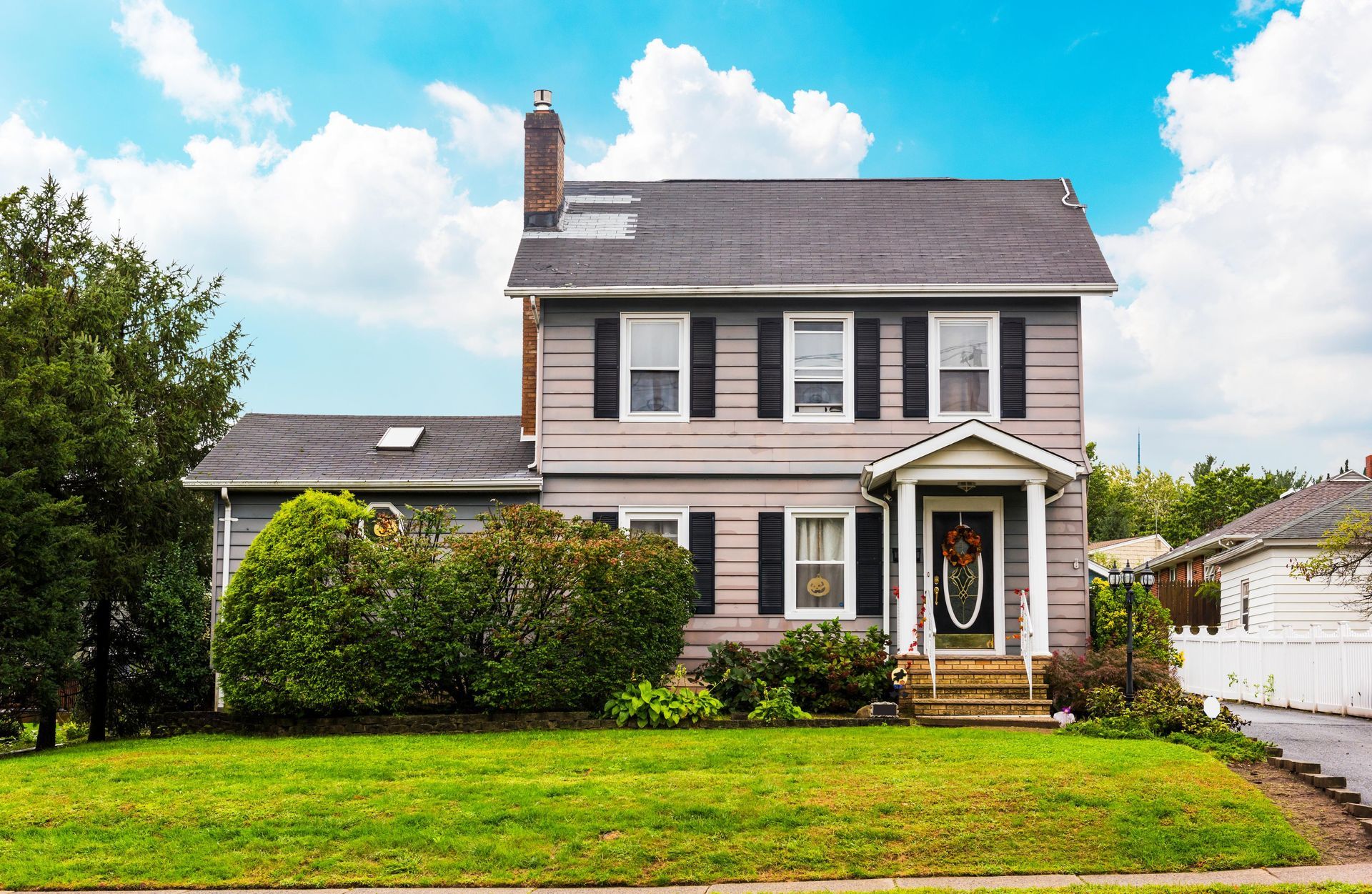 Two-story gray house with green lawn, black shutters, and front door under a small porch, blue sky in background.