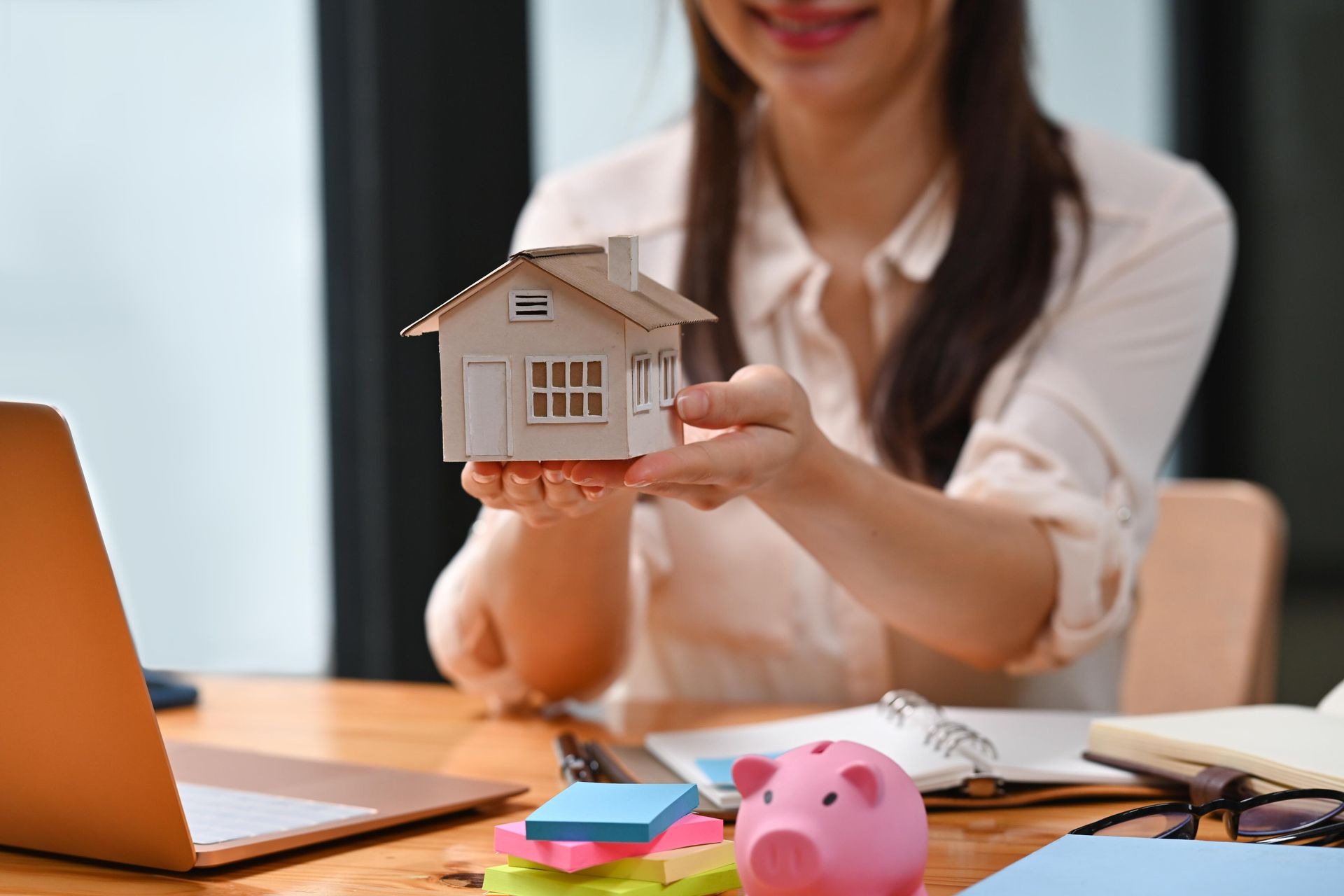 Woman holding a small house model, smiling. Desk with laptop, piggy bank, and sticky notes visible.