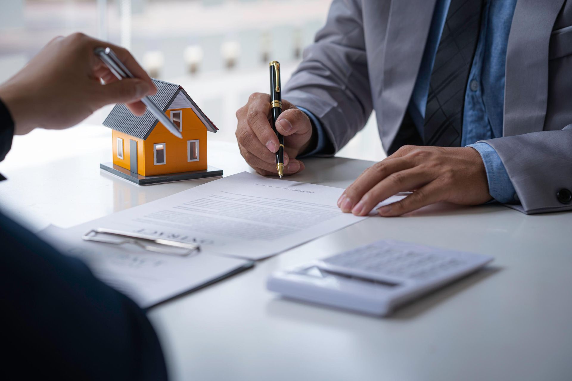 Two people signing a document on a table with a small house model and calculator.