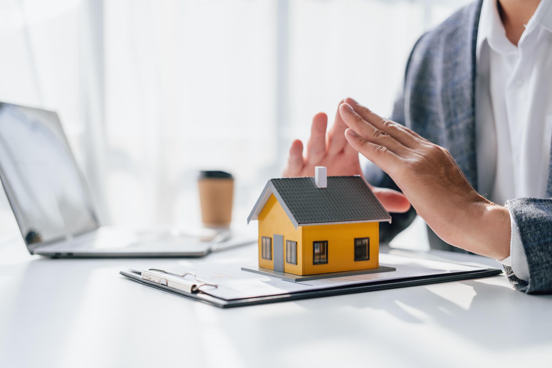 Person's hands protecting a small yellow house model on a clipboard; laptop and coffee cup nearby.