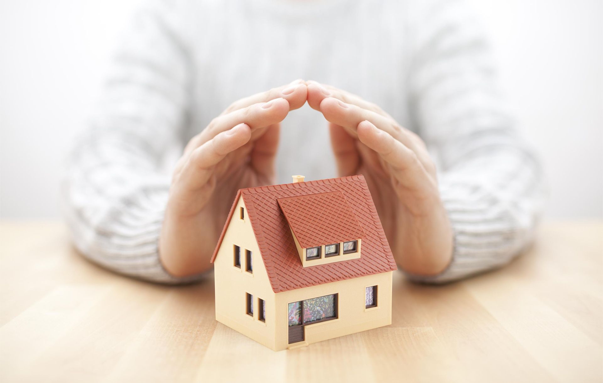 Hands cupped above a miniature house with a red roof, symbolizing protection or insurance.