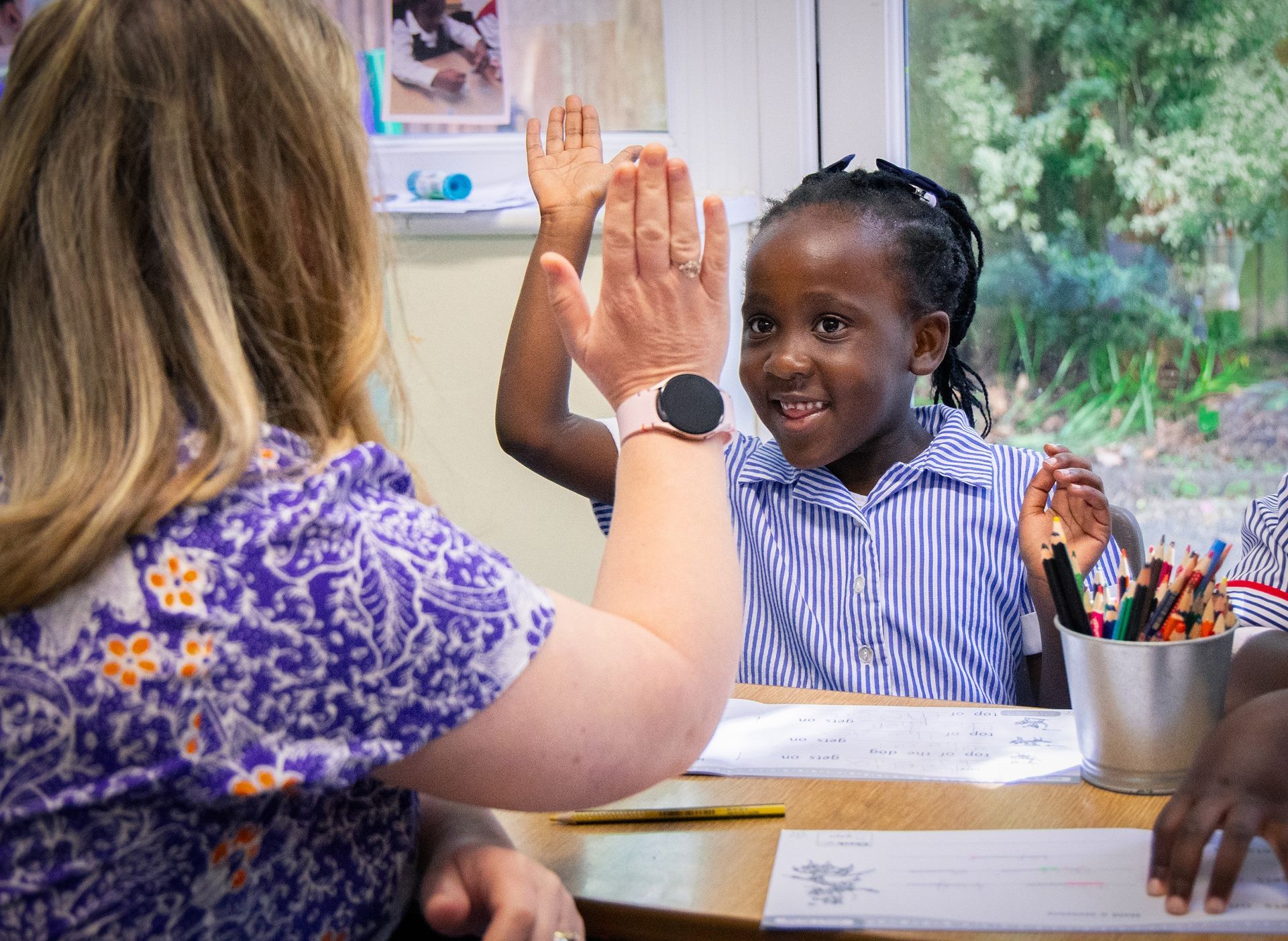 A young girl high fiving a teacher sat ata desk