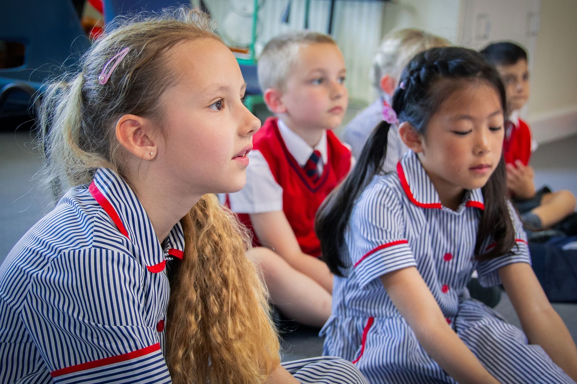 Children in striped school uniforms sitting on the floor, listening attentively in class