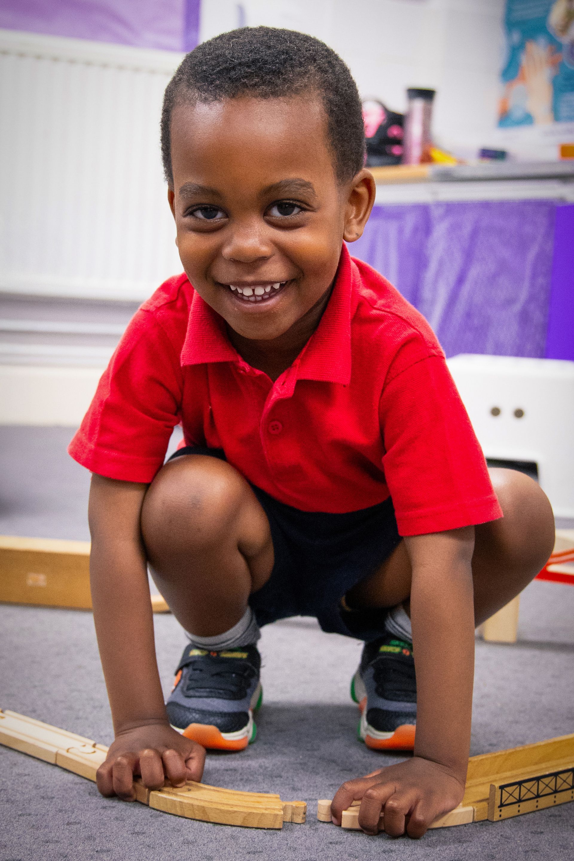 Smiling child crouches on a classroom floor, building with wooden blocks while wearing a red shirt.