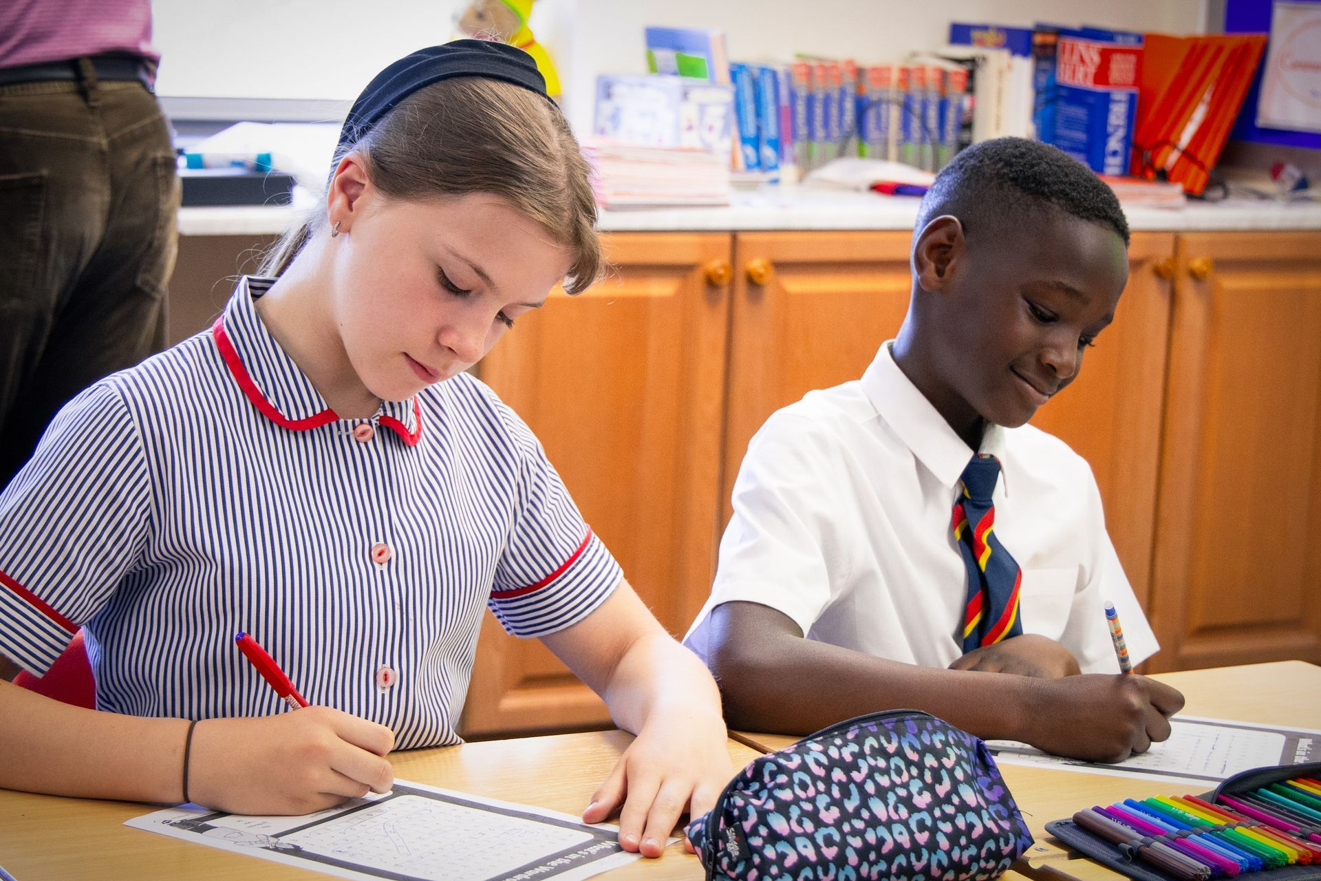 One student in a school uniform writes at a desk with colourful pencils in a classroom.