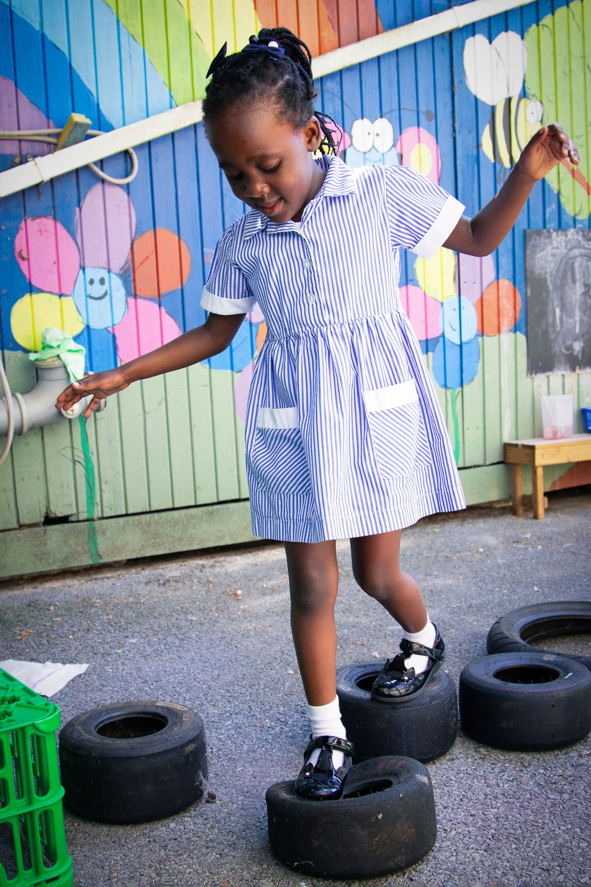 Girl balancing on tires outdoors in front of a colorful mural, arms out for balance.