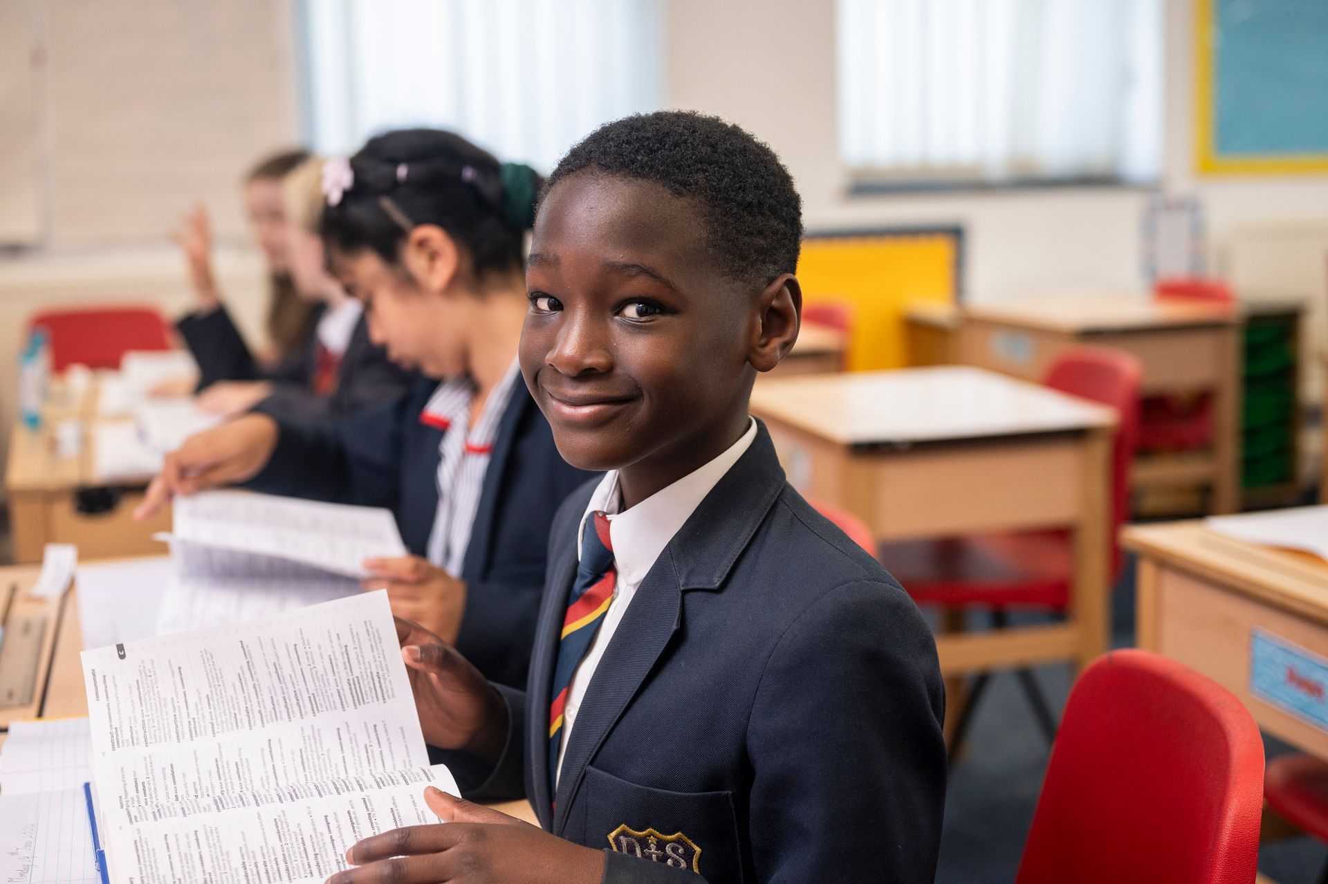 A young boy in a suit and tie is sitting at a desk in a classroom holding a piece of paper.