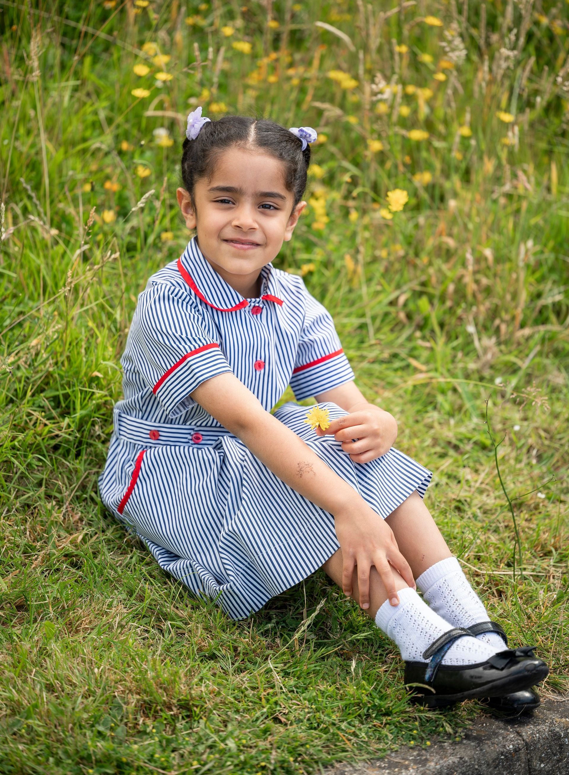 A little girl in a school uniform is sitting in the grass holding a flower.
