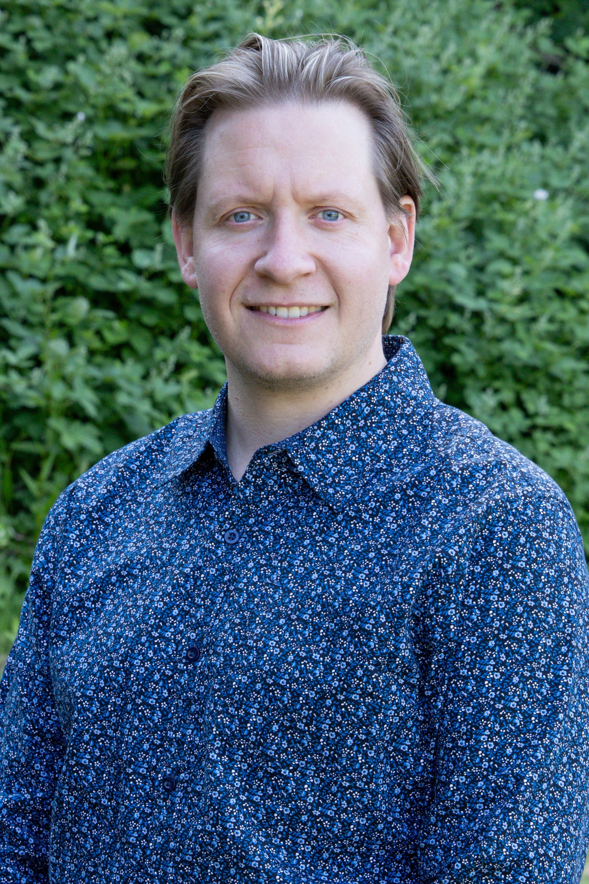 A man in a blue shirt is smiling for the camera in front of a bush.
