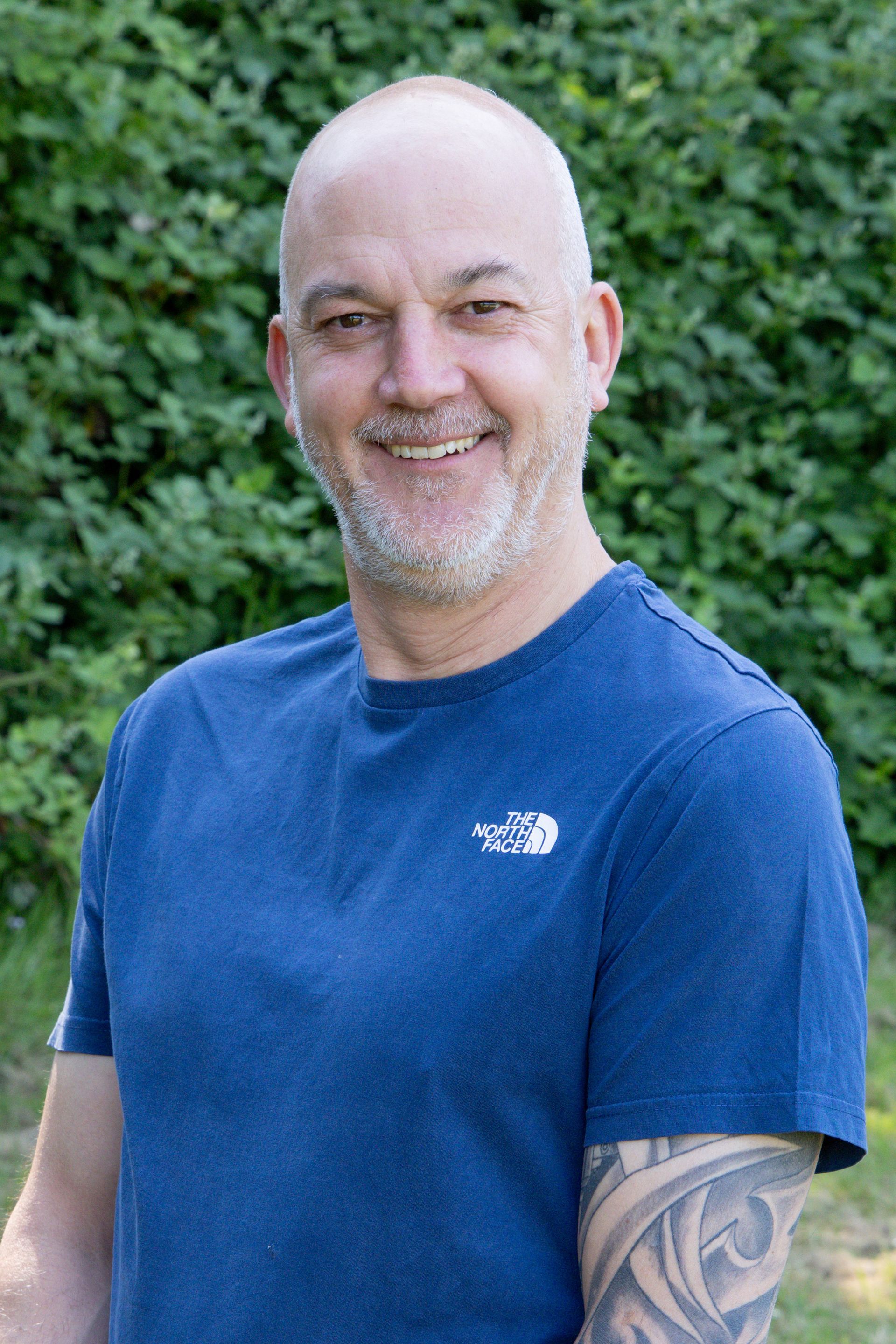 A man with a tattoo on his arm is wearing a blue shirt and smiling.