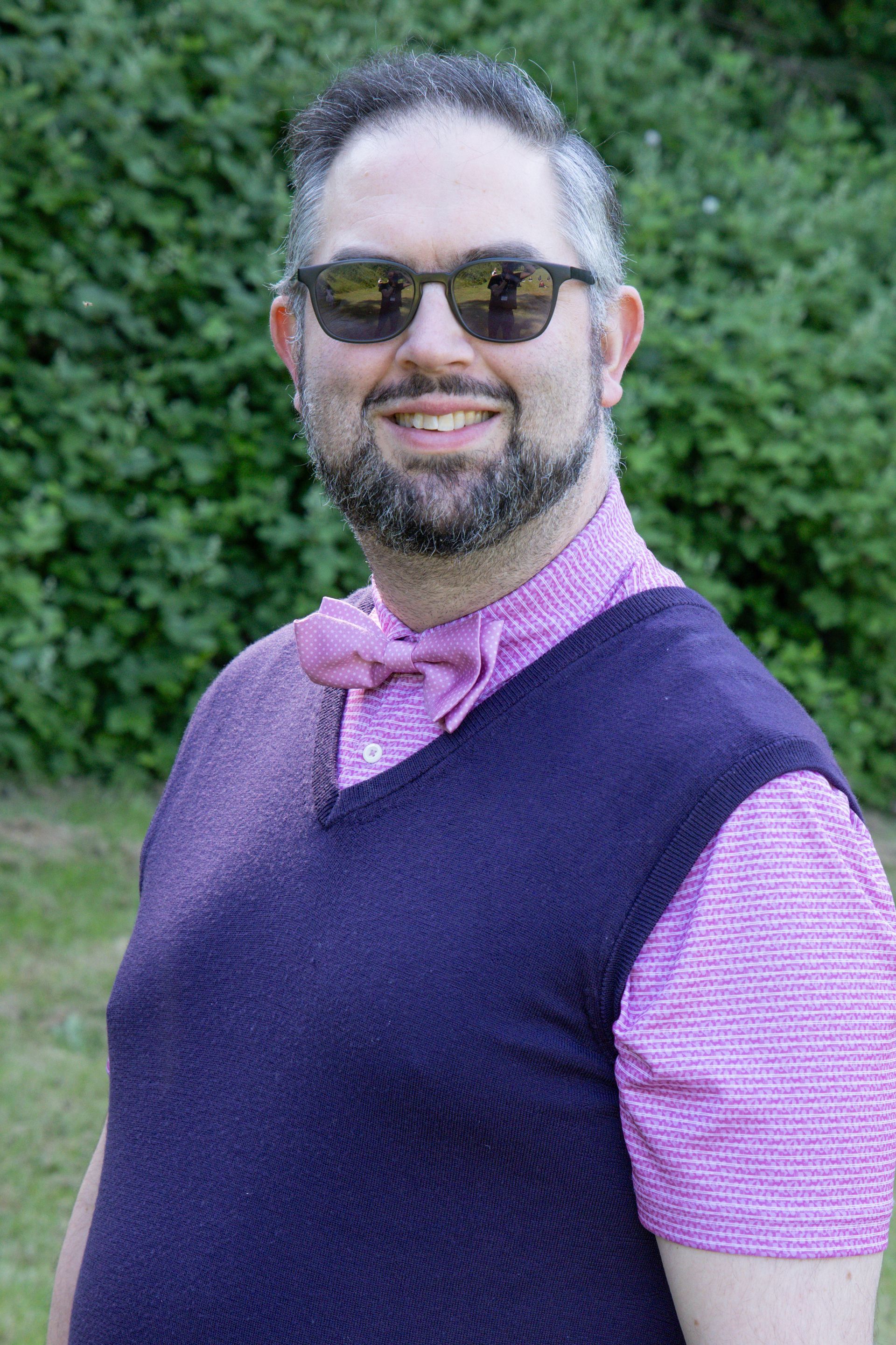A man with a beard and sunglasses is wearing a purple vest and a pink shirt.