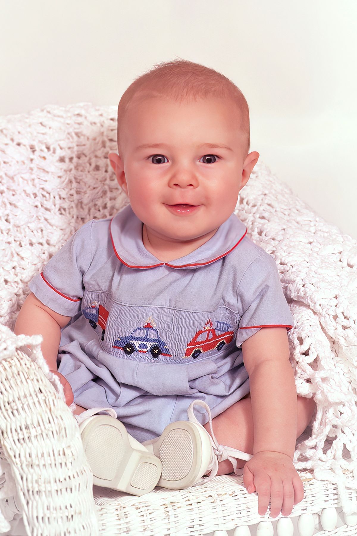 A baby is sitting on a white blanket wearing a fire truck outfit.