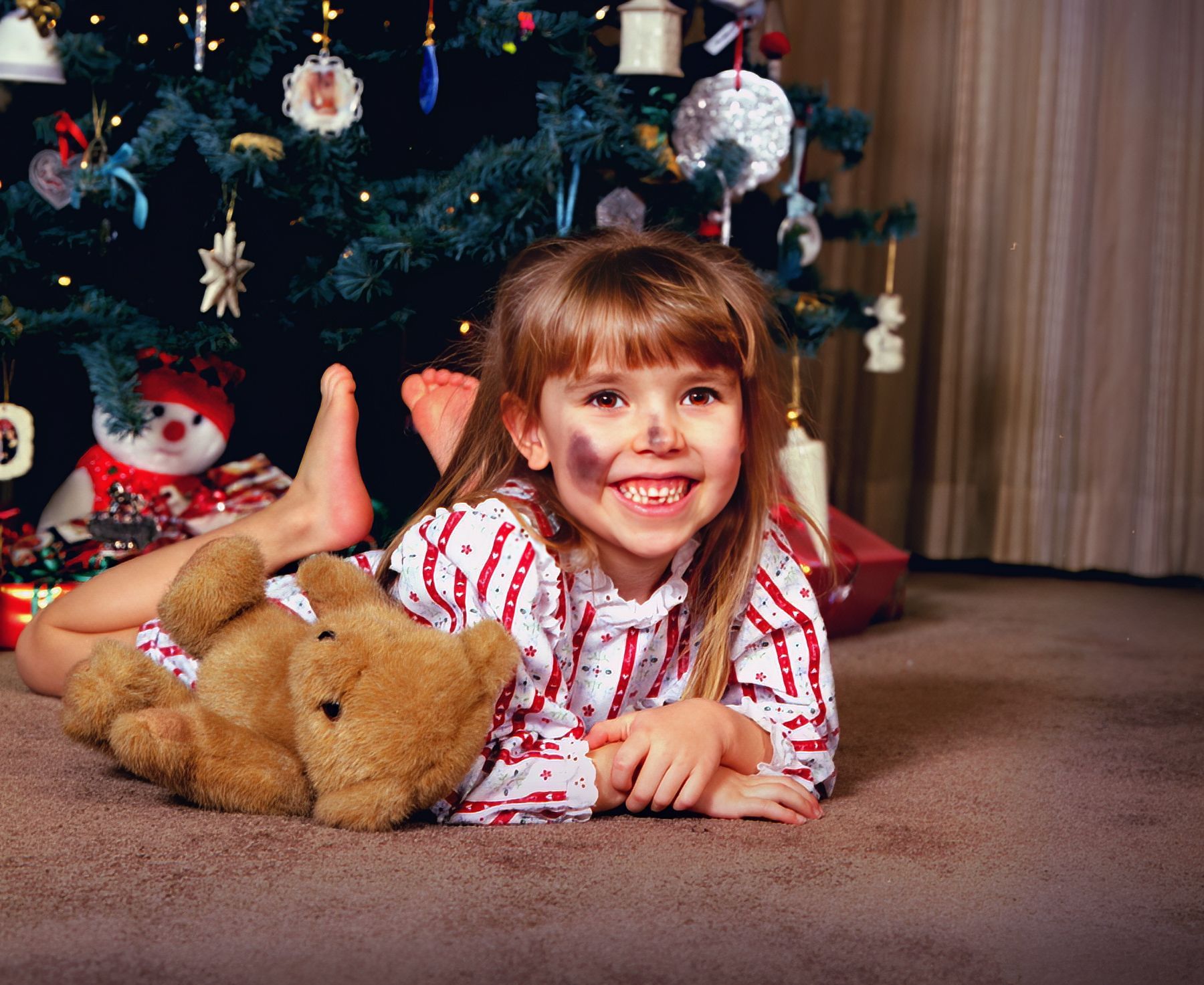 A little girl laying on the floor with a teddy bear in front of a christmas tree