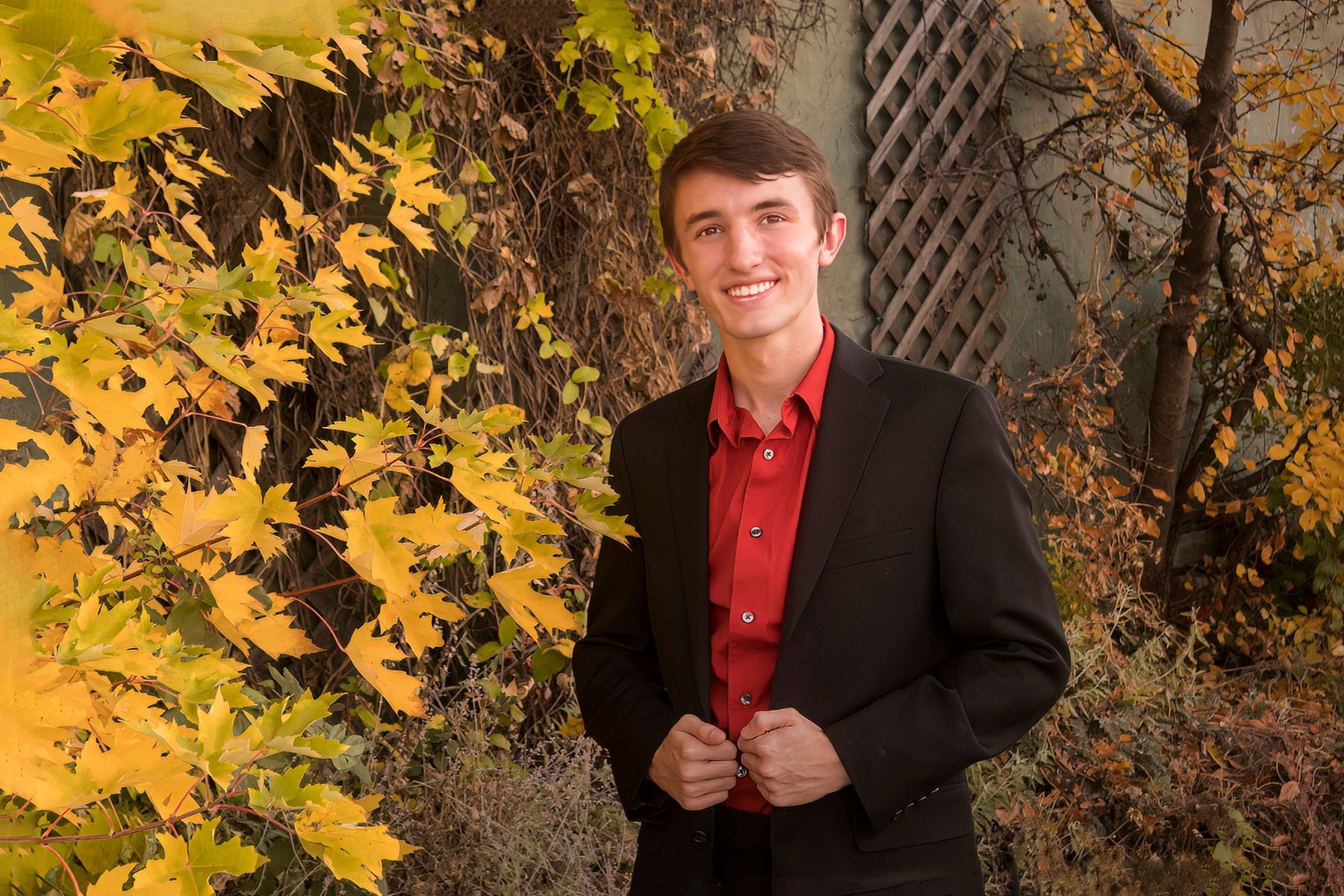 A young man in a suit and red shirt is standing in front of a tree.