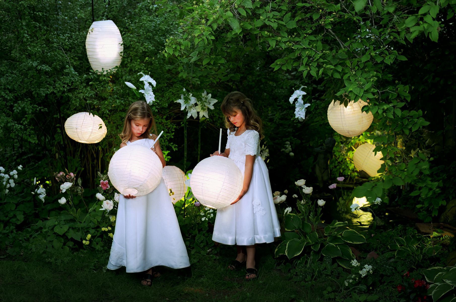 Two little girls in white dresses are holding lanterns in a garden.