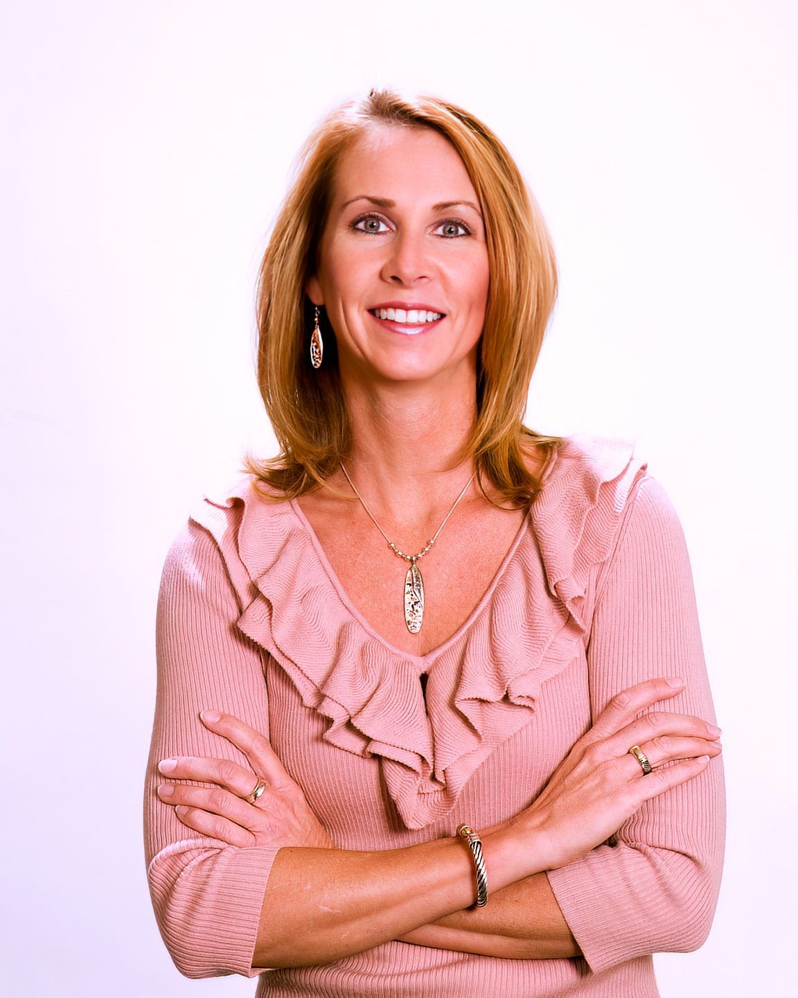 A woman in a pink ruffled top is smiling with her arms crossed