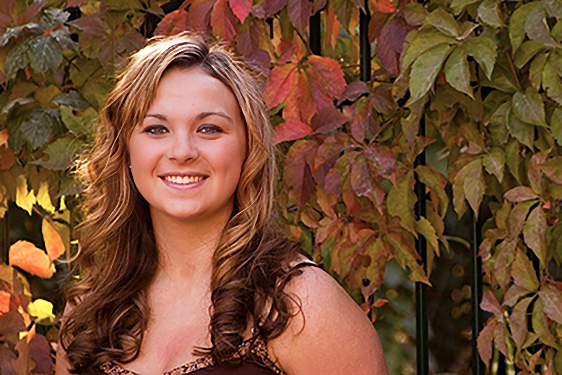 A woman is smiling in front of a fence with leaves in the background