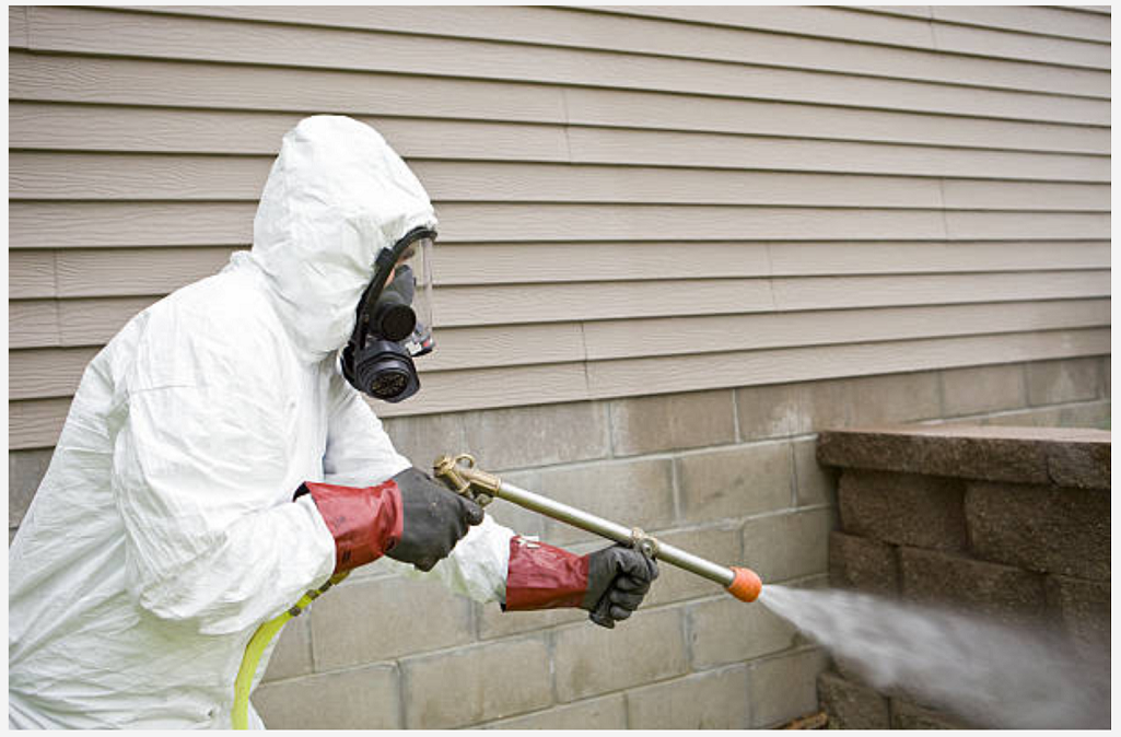 Un uomo con una tuta protettiva sta spruzzando una casa con uno spruzzatore.