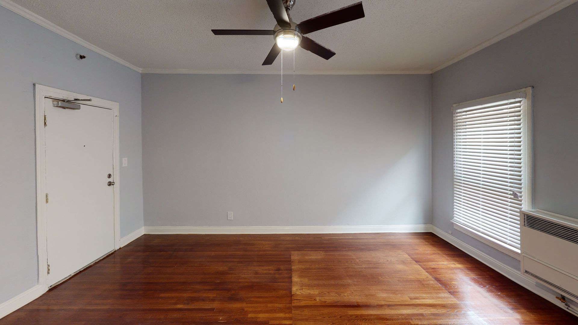 Empty room with hardwood floors, light blue walls, door, window with blinds, and ceiling fan.